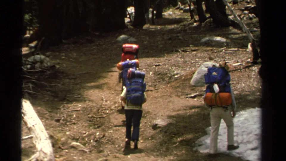 1978 Lake Tahoe frame showing backpackers moving deeper into a shaded forest trail with snow patches and rocks nearby.