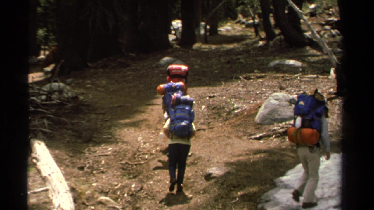 Two hikers with large backpacks walk through a Lake Tahoe forest trail in 1978 home-movie footage, with trees and snow patches visible.