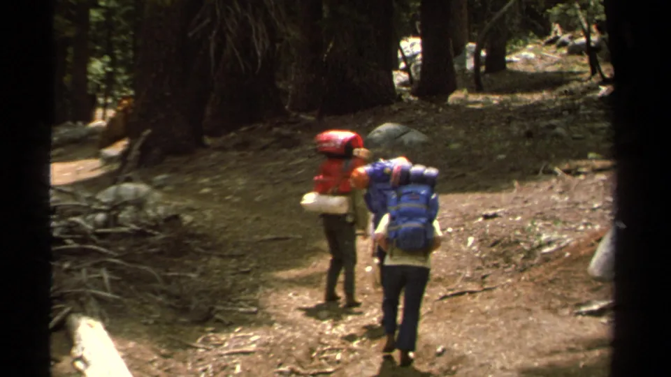 Two backpacked hikers move through a Lake Tahoe forest in 1978 home-movie footage, framed by dark trunks and uneven trail ground.