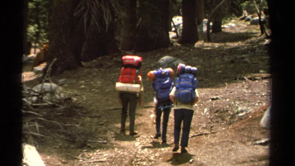 Three hikers with colorful backpacks walk away on a Lake Tahoe forest trail in 1978 8mm home-movie footage.