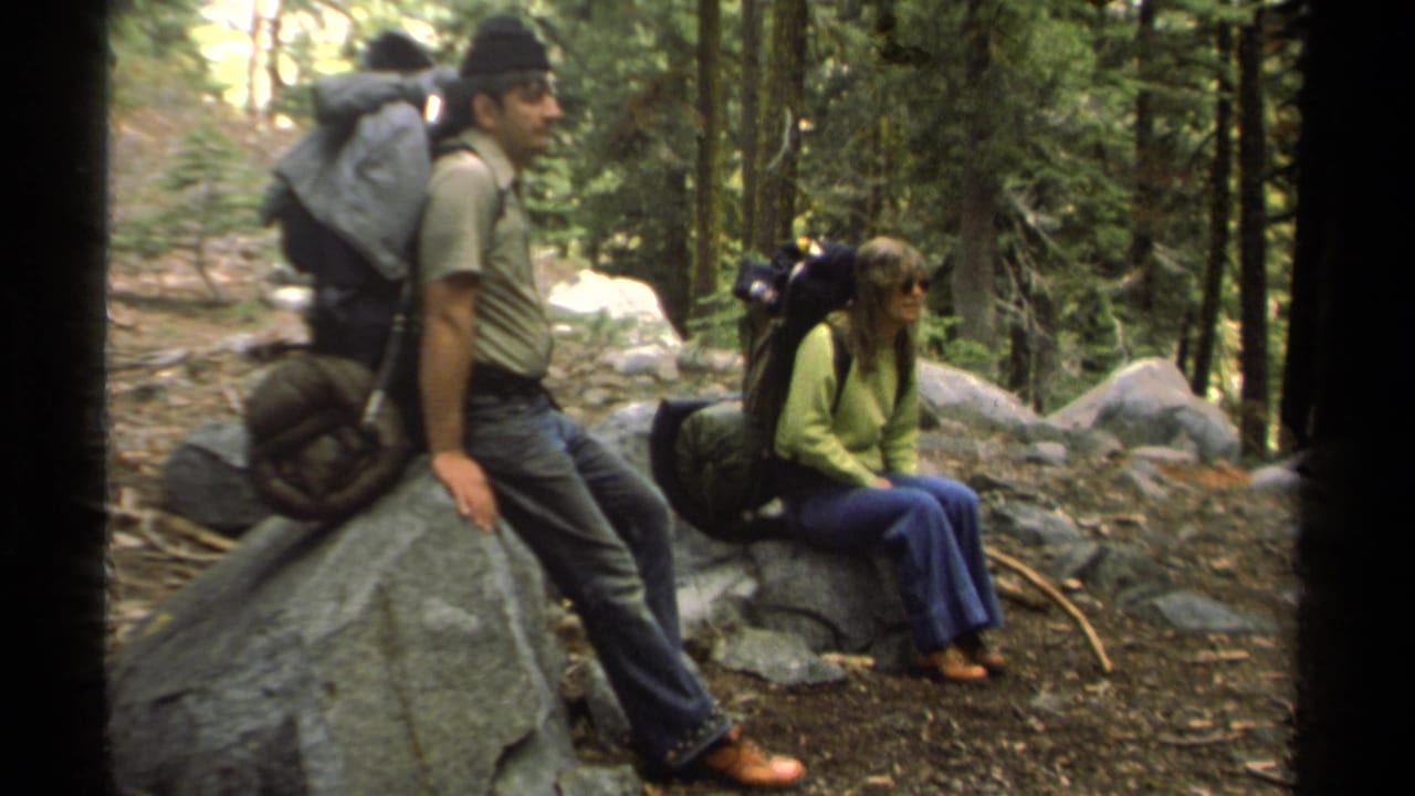 A man and woman with camping packs pause on rocks in a Lake Tahoe pine forest, filmed on 8mm in 1978.
