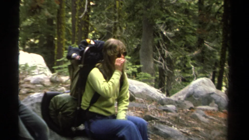 A woman wearing sunglasses and a large pack sits on a forest rock in 1978 Lake Tahoe home-movie footage.