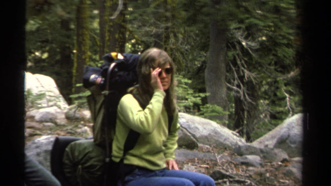 In 1978 Lake Tahoe footage, a woman in a green sweater and blue jeans rests on a rock with a large backpack and sunglasses.