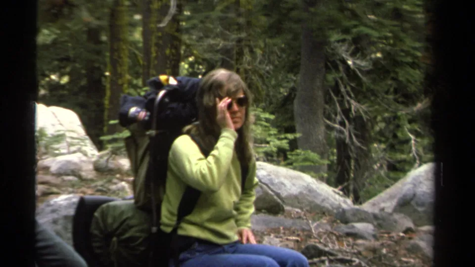 In 1978 Lake Tahoe footage, a woman in a green sweater and blue jeans rests on a rock with a large backpack and sunglasses.