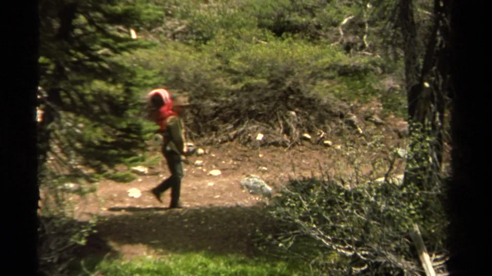 A distant hiker with a red backpack walks along a dirt forest trail in the Lake Tahoe 1978 home-movie scene.
