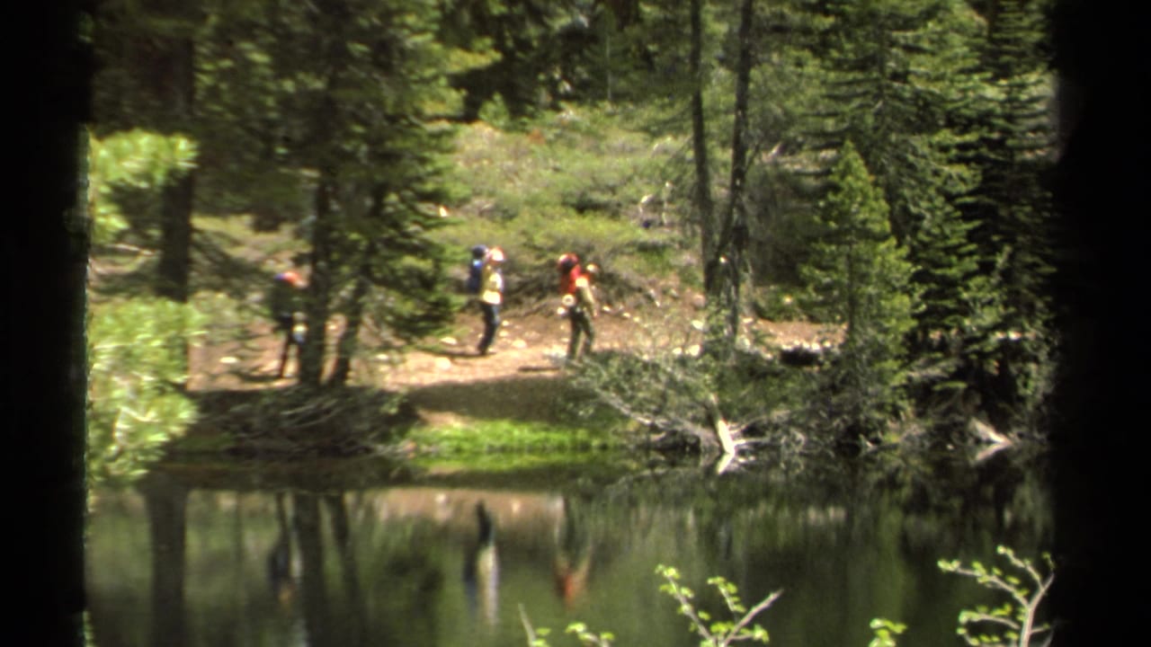 Several distant hikers with backpacks walk along a forested Lake Tahoe shoreline path, reflected faintly in the still water.