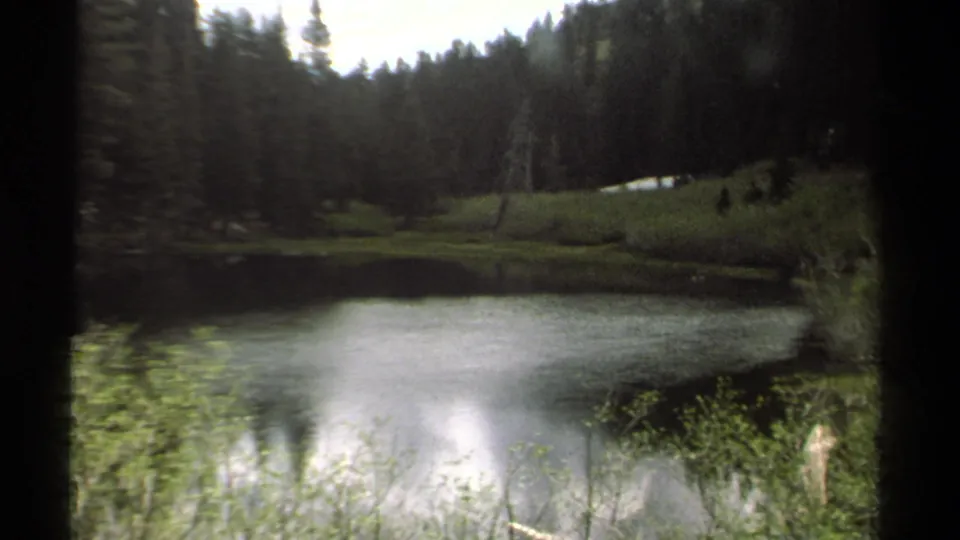 1978 Lake Tahoe 8mm view of a quiet forest lake, with soft exposure, dark borders, and low vegetation along the near shore.
