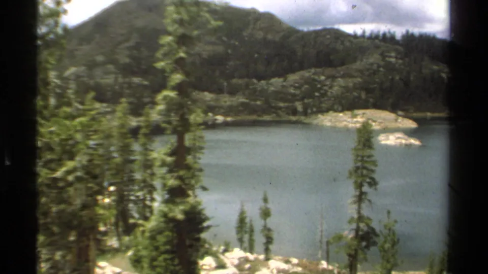1978 Lake Tahoe 8mm frame showing blue-green water, conifers, a rocky shoreline, and a wooded slope in the distance.