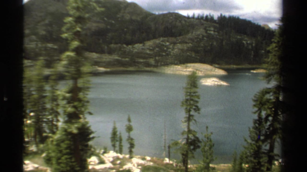 1978 8mm Lake Tahoe view of a blue-green mountain lake framed by evergreens, rocky islands, wooded hills, and cloudy sky.