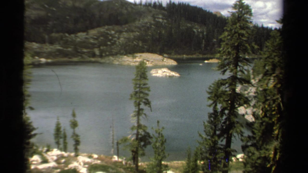 1978 Lake Tahoe 8mm frame showing calm lake water, foreground pines, pale rock outcrops, and dark evergreen hillsides.