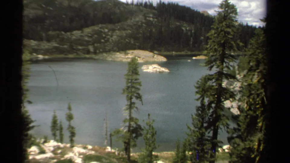 1978 Lake Tahoe 8mm frame showing calm lake water, foreground pines, pale rock outcrops, and dark evergreen hillsides.