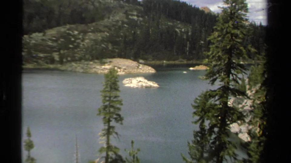 1978 8mm Lake Tahoe view with a closer framing of rocky islands, pine trees, lake water, and a forested slope behind.
