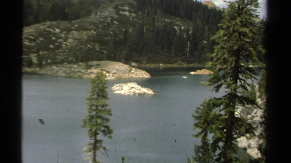 1978 Lake Tahoe home-movie frame with a zoomed composition of a rocky island, blue-green water, pines, and wooded hills.