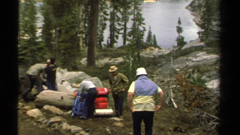 A small group of campers with trail gear stands near boulders and trees above Lake Tahoe in 1978 8mm footage.