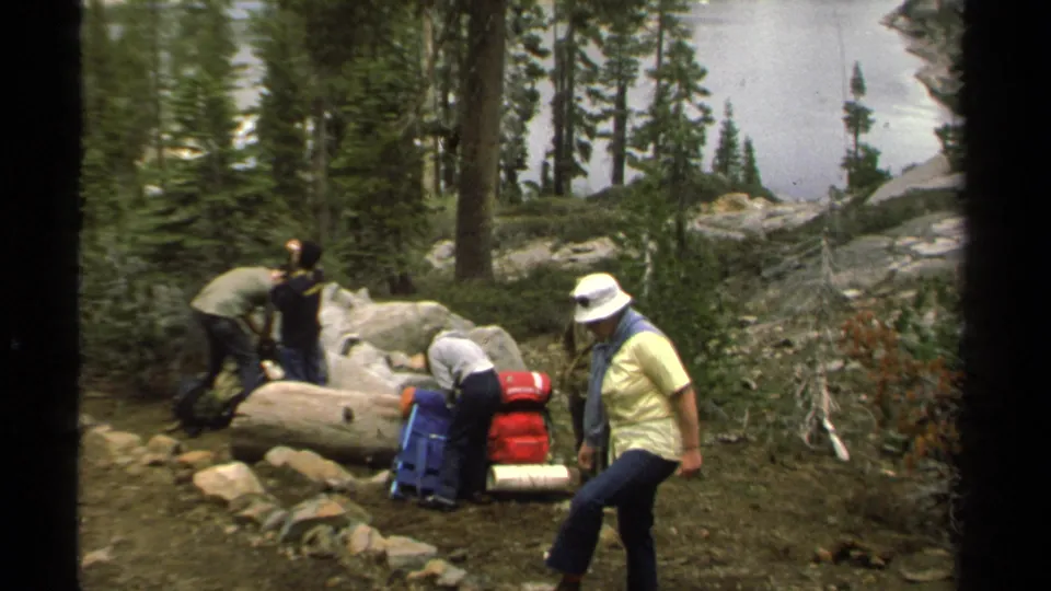 Lake Tahoe campers in casual trail clothing move around packed gear beside rocks and pine trees in a 1978 home movie.