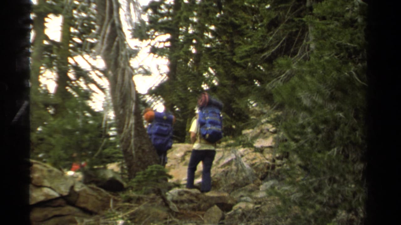 Two backpackers walk away through trees and rocks on a Lake Tahoe trail, filmed on 8mm in 1978.