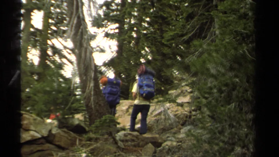 Two backpackers walk away through trees and rocks on a Lake Tahoe trail, filmed on 8mm in 1978.