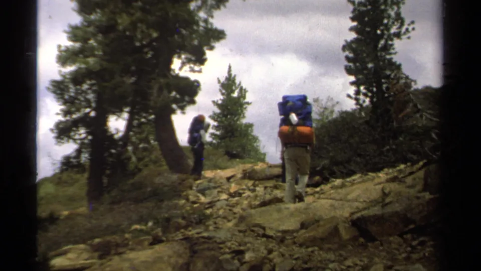 A hiker with a blue pack and orange roll follows another person on a rocky Lake Tahoe forest trail in 1978.