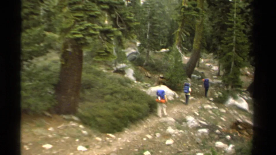 Three backpackers walk through a pine forest trail in the Lake Tahoe area, seen from a handheld 1978 8mm camera.