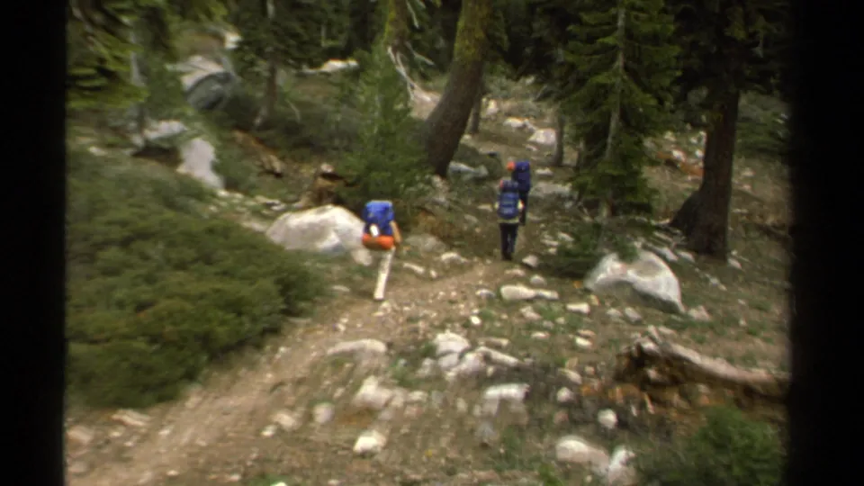 Three hikers with blue backpacks move along a rocky wooded trail in Lake Tahoe, California, in 1978 home-movie footage.