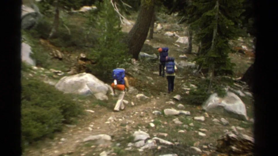 Backpackers continue single file on a rocky Lake Tahoe forest trail, seen from behind in 1978 8mm footage.