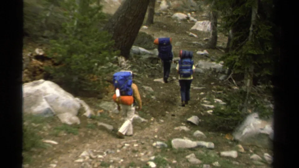 Three backpackers walk away from the camera through a shaded rocky Lake Tahoe forest path in archival 8mm footage.
