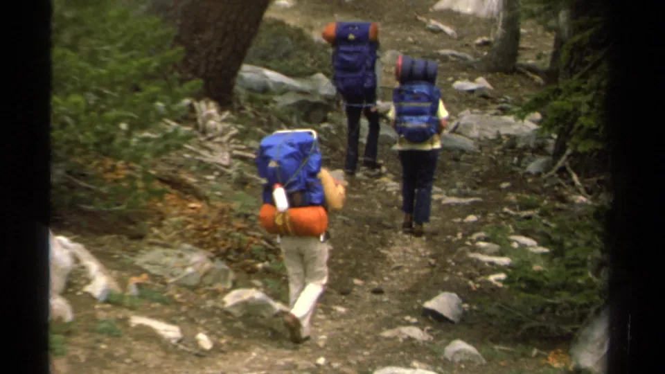Two foreground hikers with large blue backpacks follow another hiker down a rocky Lake Tahoe trail in 1978 home-movie footage.