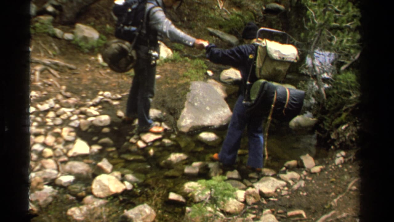 Backpackers pause at a shallow rocky stream in the Lake Tahoe woods, with one hiker offering help.