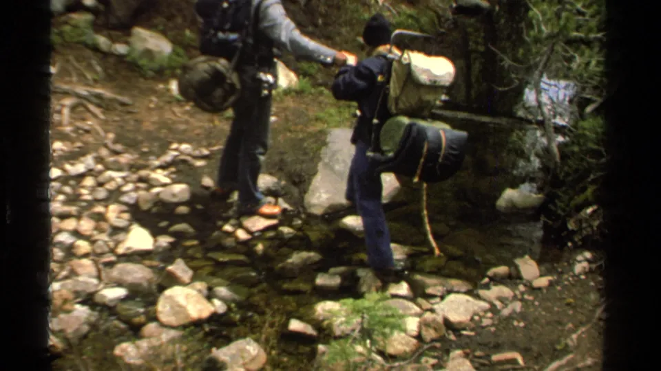 A backpacker balances on rocks in a shallow Lake Tahoe stream as another hiker stands close by.