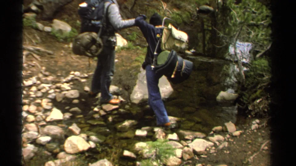 In 1978 Lake Tahoe home-movie footage, one backpacker steadies another crossing a shallow rocky stream.