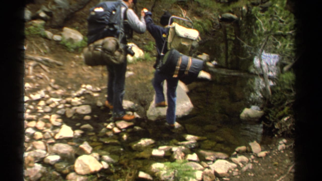 1978 Lake Tahoe home-movie frame showing two hikers with large packs crossing rocks over a shallow forest stream.