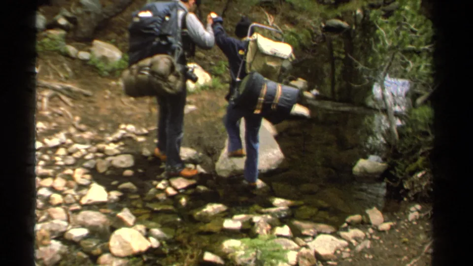 1978 Lake Tahoe home-movie frame showing two hikers with large packs crossing rocks over a shallow forest stream.