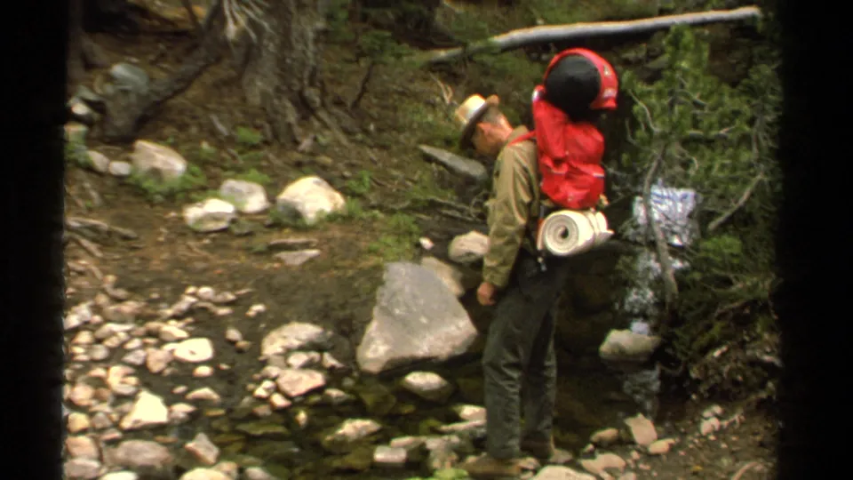 1978 Lake Tahoe frame showing a hiker with a red pack and straw hat paused beside a shallow forest stream.