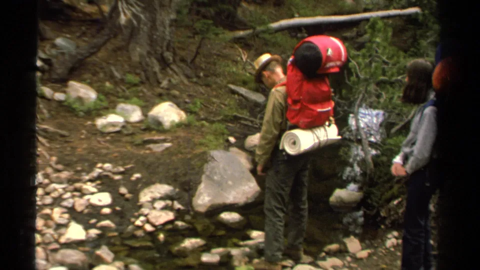 Lake Tahoe 1978 home-movie frame with a red-pack hiker near a rocky stream and another hiker at the edge of the shot.