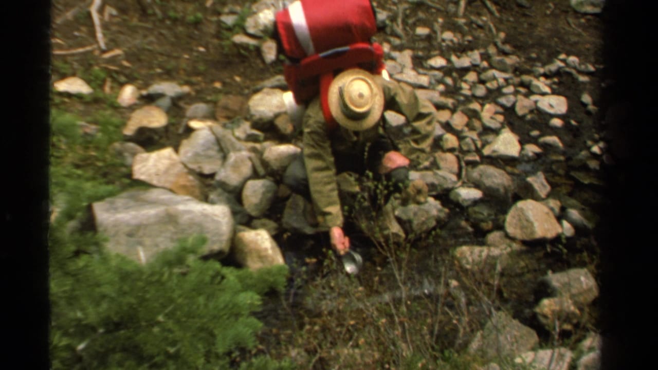 1978 Lake Tahoe home-movie frame of a hiker crouching among rocks near a stream, wearing a straw hat, olive jacket, and red pack.