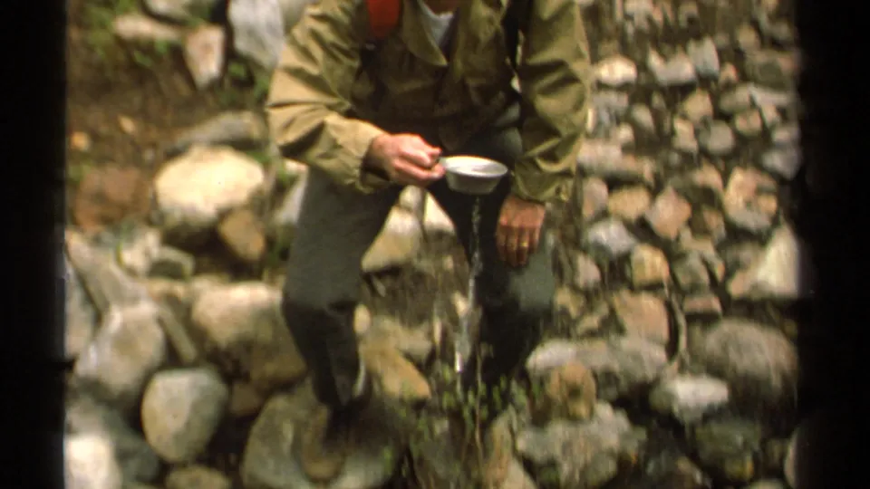 Soft 8mm close frame shows the hiker holding a metal cup after scooping water from the rocky stream.