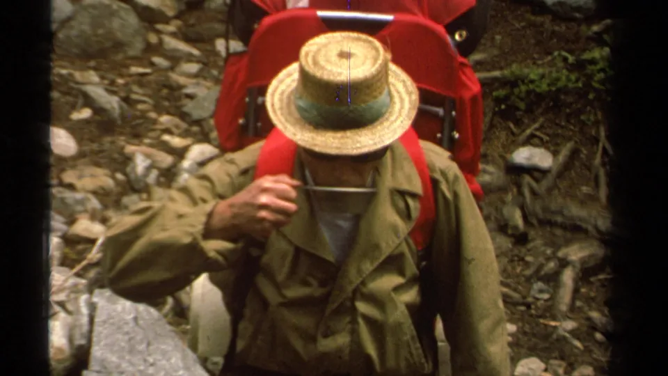 1978 Lake Tahoe home-movie frame showing a man in a straw hat and olive jacket on a rocky mountain trail with red hiking gear.