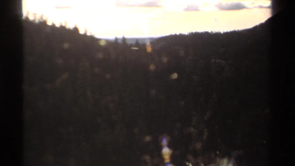 1978 home-movie landscape frame showing a shadowed mountain forest valley below a bright sky, softened by grain and exposure bloom.