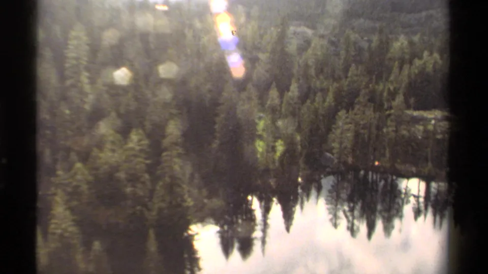 Lake Tahoe area 8mm frame of a calm lake reflecting surrounding pine trees and hillside, with strong sunlight and visible lens flare.