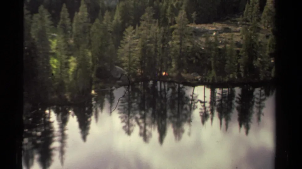 Closer 1978 Lake Tahoe frame showing a small tent tucked into evergreens beside reflective lake water.