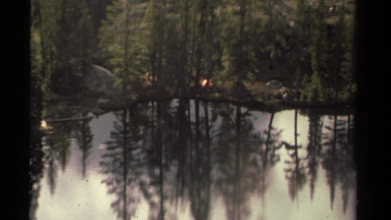 1978 8mm Lake Tahoe-area view of calm water reflecting pine forest, with a small campfire glow across the lake.