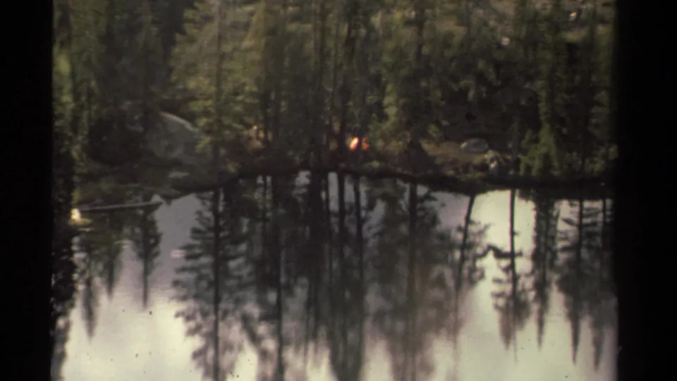 1978 8mm Lake Tahoe-area view of calm water reflecting pine forest, with a small campfire glow across the lake.