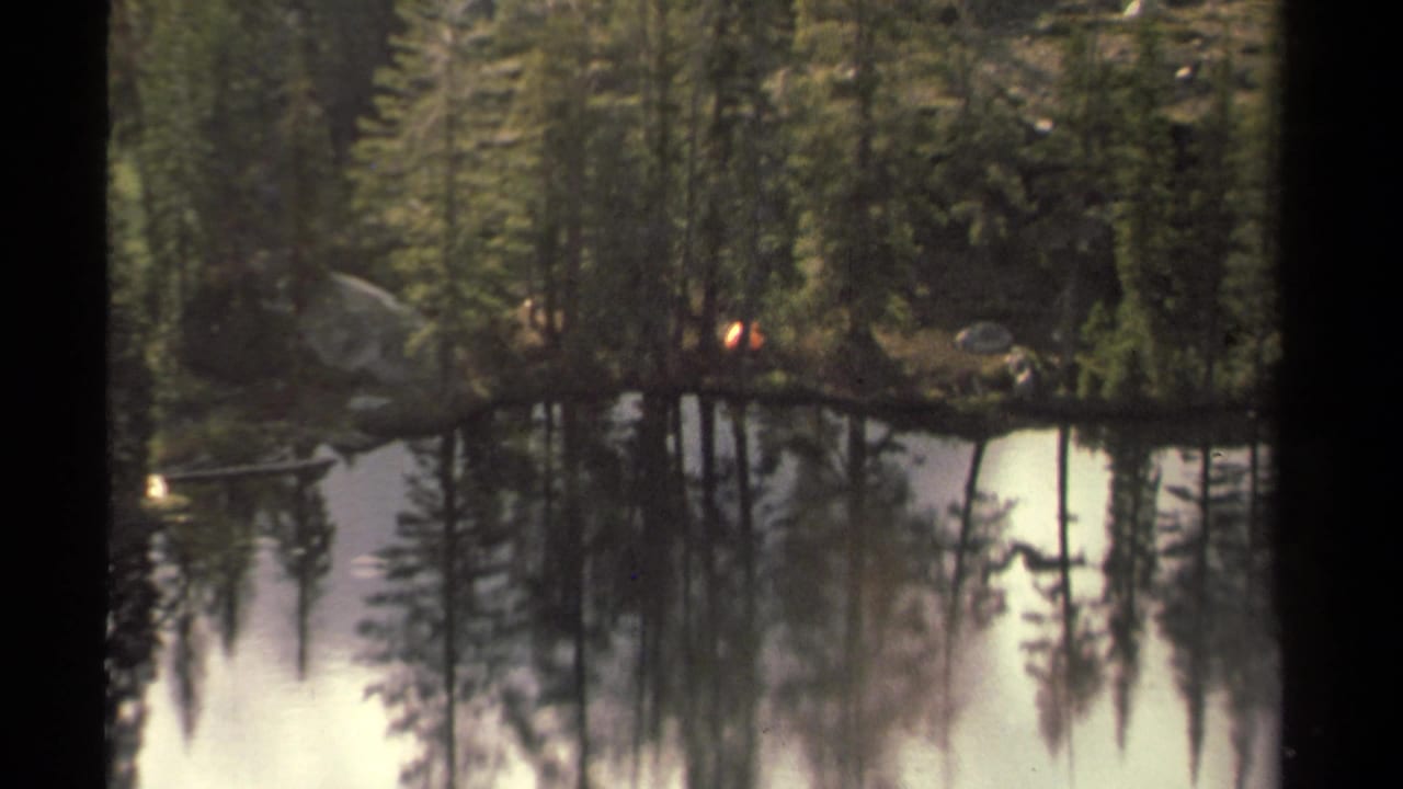 Lake Tahoe-area forest lake scene on 8mm film, showing reflected pines and a small campfire point near the far bank.