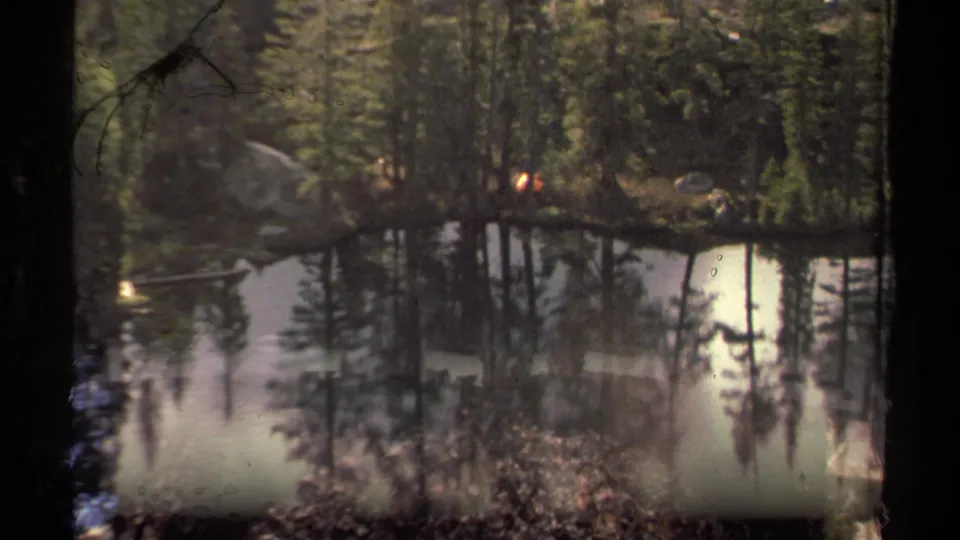 Vintage 8mm view of Round Lake area water and pine reflections, with a small campfire glow visible across the lake.