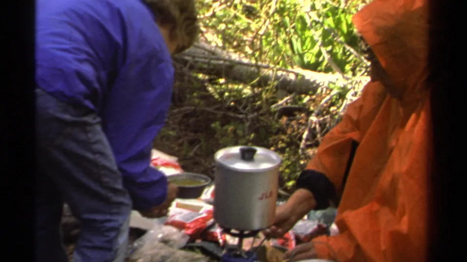 In wooded Lake Tahoe camping footage from 1978, a silver pot on a gas stove is surrounded by food packets and rain gear.