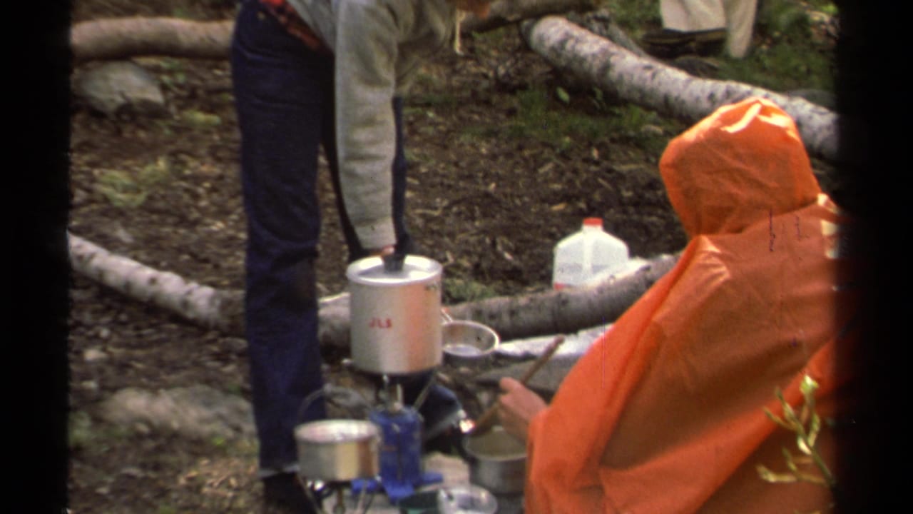 1978 Lake Tahoe home-movie frame showing campers around a blue camp stove, silver cookware, a plastic water jug, and an orange poncho.