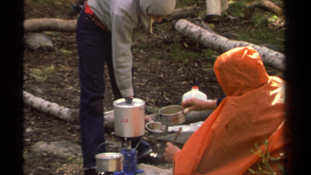 1978 Lake Tahoe camping frame with a seated person in an orange poncho holding a metal cup near boiling cookware and a water jug.