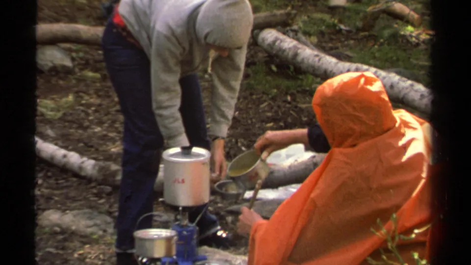 1978 Lake Tahoe home-movie frame of a hooded camper tending a camp stove as a seated person in an orange poncho holds a metal cup.