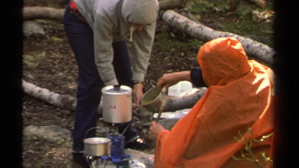 1978 Lake Tahoe camping frame showing simple outdoor cooking gear, a hooded person, and a seated camper in an orange poncho.