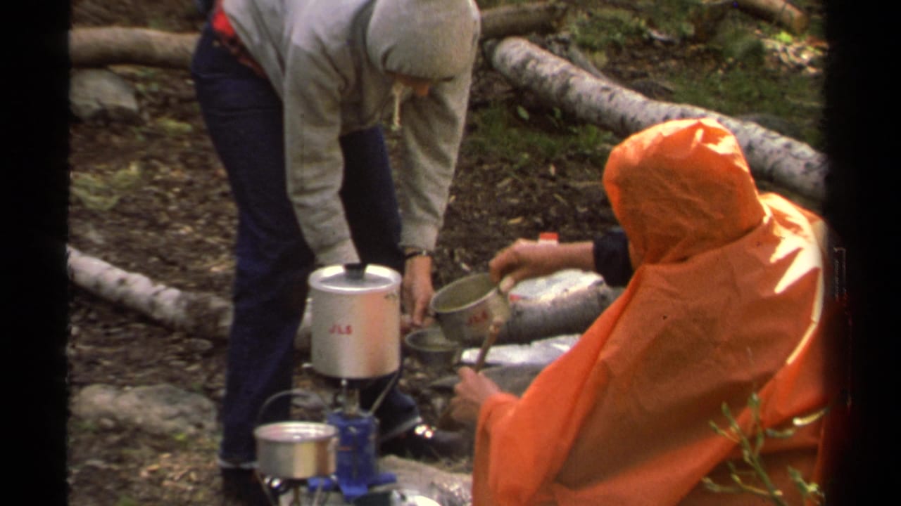 Two people prepare food beside a portable camp stove in a wooded area.